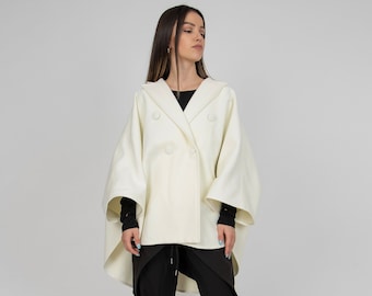 May include: An off-white double-breasted cape jacket with a wide collar and four matching buttons, worn over a black long-sleeved top and black trousers with a drawstring waist, paired with black open-toe ankle boots with a block heel.