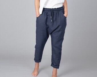May include: A woman wearing a white tank top and dark blue linen pants with a drawstring waist. She is standing on a gray background.