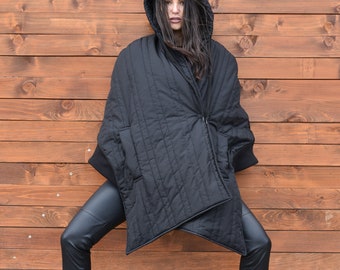 May include: A woman wearing a black hooded quilted jacket with a zipper and black leather pants. She is standing in front of a wooden wall.