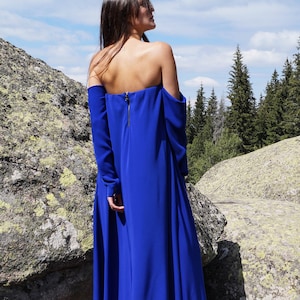 Könnte beinhalten: Ein langes, königsblaues schulterfreies Kleid mit langen Ärmeln. Das Kleid hat einen goldenen Reißverschluss am Rücken und einen fließenden Rock. Das Model steht auf einer Felsformation mit Wald und blauem Himmel im Hintergrund.
