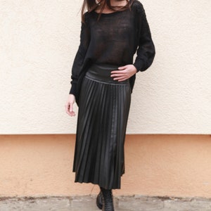 May include: A woman wearing a black wide-brimmed hat, a black sweater, and a black pleated skirt. She is standing in front of a wall and looking down.