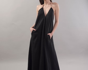 May include: A woman wearing a black maxi dress with a plunging neckline and thin straps. She is wearing sunglasses and has her hands in her pockets.
