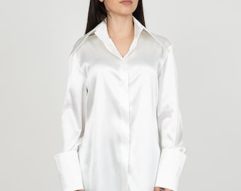 May include: A white satin shirt with a long sleeve and a button-up front. The shirt has a collar and a relaxed fit.