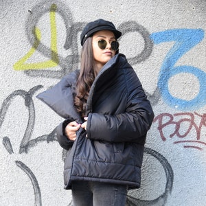 May include: A black puffer jacket with a large collar and a zipper closure. The jacket is worn by a person with long brown hair and sunglasses. The person is standing in front of a wall with graffiti.