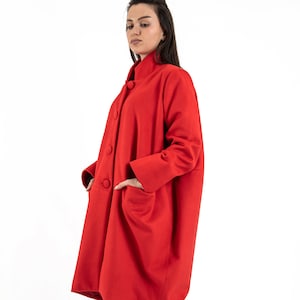 Könnte beinhalten: Ein langärmliger roter Mantel mit Stehkragen und Knopfleiste. Der Mantel hat große Taschen vorne und wird über einer blauen Jeans und schwarzen Sneakern getragen.