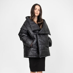 May include: A black puffer jacket with a large, asymmetrical collar. The jacket is quilted and has a shiny finish. The model is wearing a black skirt and clear heels.