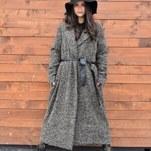 Puede incluir: Una mujer con un abrigo largo de tweed gris y negro con un cinturón de cuero negro. También lleva un sombrero negro y botas negras.