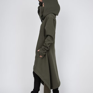 May include: An olive green hooded coat with a high collar and a long, asymmetrical hem. The coat features a zipper closure and thumbhole cuffs. The person is wearing black pants and high-heeled boots.