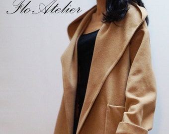 May include: A camel-colored hooded coat featuring a wide lapel, long sleeves with folded cuffs, and two large patch pockets. The coat is open at the front, revealing a black top and dark blue jeans. The text Flo Atelier is visible in the top left corner.