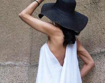 May include: A white linen dress with a draped neckline and a wide, flowing skirt. The dress is worn by a person wearing a black wide-brimmed hat.