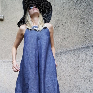 May include: A floor-length blue dress with a high neckline and a thick, off-white rope detail around the collar. The dress features a loose, flowing silhouette. A wide-brimmed black hat with white polka dots and dark sunglasses are also worn.