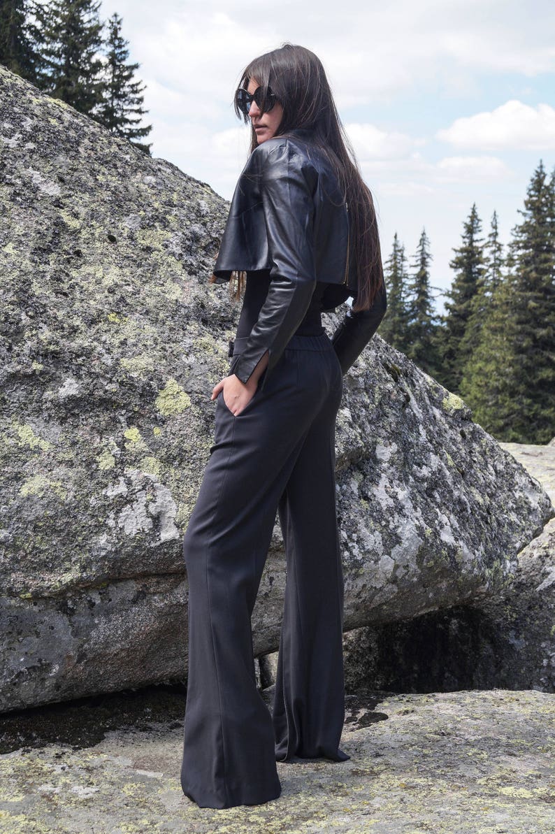 May include: A woman wearing a black leather jacket with a cropped design and black wide-leg pants. She is standing in front of a large rock with a forest in the background.