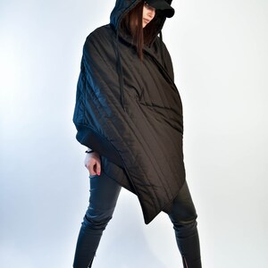 May include: A black quilted hooded jacket with a long, asymmetrical hemline. The jacket has a zipper closure and a drawstring hood. The model is wearing black pants and sneakers.