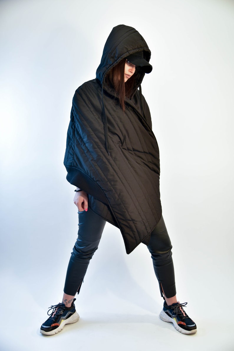 May include: A black quilted hooded jacket with a long, asymmetrical hemline. The jacket has a zipper closure and a drawstring hood. The model is wearing black pants and sneakers.