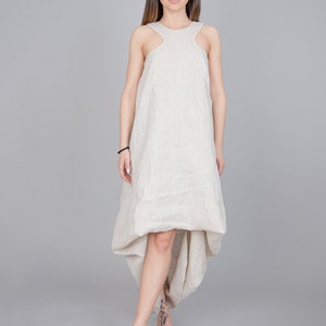 May include: A woman wearing a beige linen dress with a high neckline and a high-low hemline. The dress has a relaxed fit and is sleeveless. The woman is standing on a gray background.