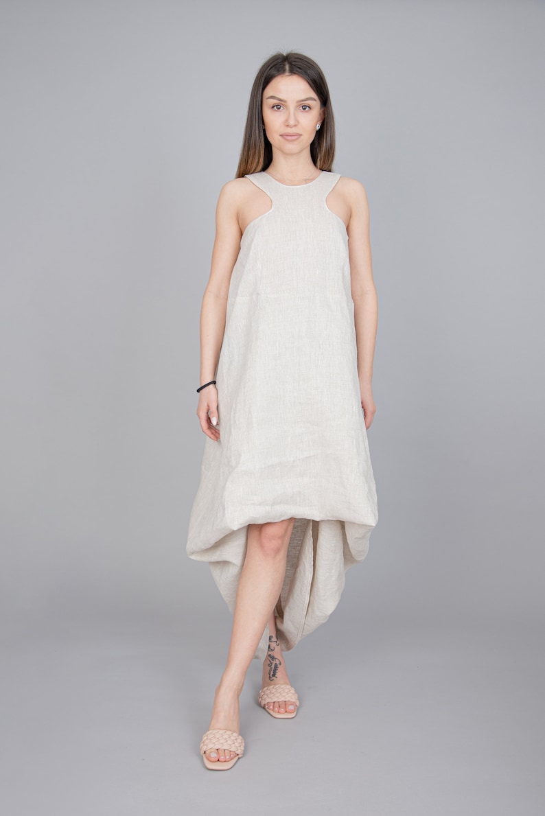 May include: A woman wearing a beige linen dress with a high neckline and a high-low hemline. The dress has a relaxed fit and is sleeveless. The woman is standing on a gray background.