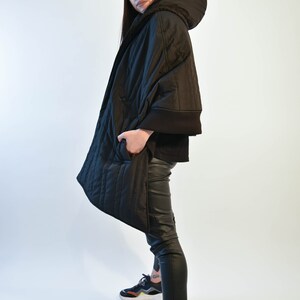May include: A black quilted jacket with a hood and a black baseball cap. The jacket has a zipper closure and pockets. The person is wearing black pants and black sneakers.