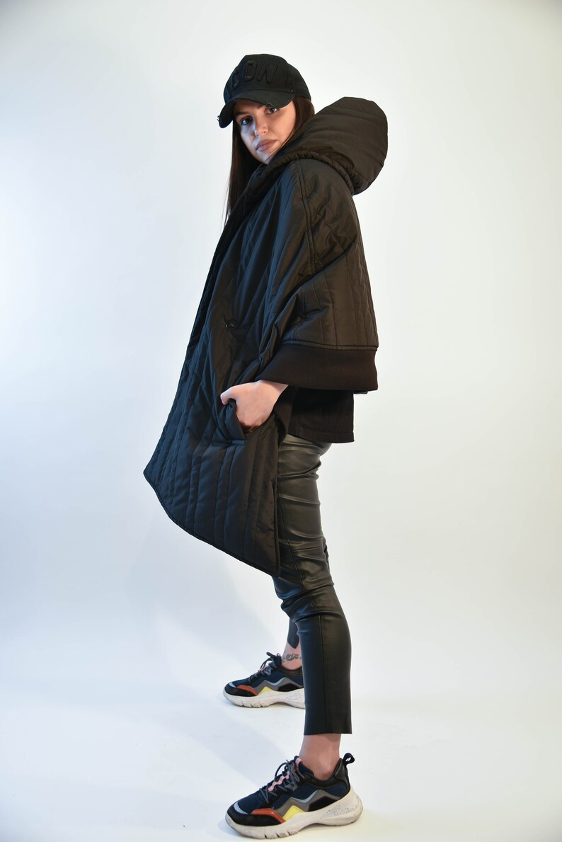 May include: A black quilted jacket with a hood and a black baseball cap. The jacket has a zipper closure and pockets. The person is wearing black pants and black sneakers.