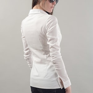 Puede incluir: Una camisa blanca de manga larga con botones y silueta ajustada. La camisa tiene cuello y mangas largas con pu&ntilde;os abotonados. La modelo lleva pantalones vaqueros negros.