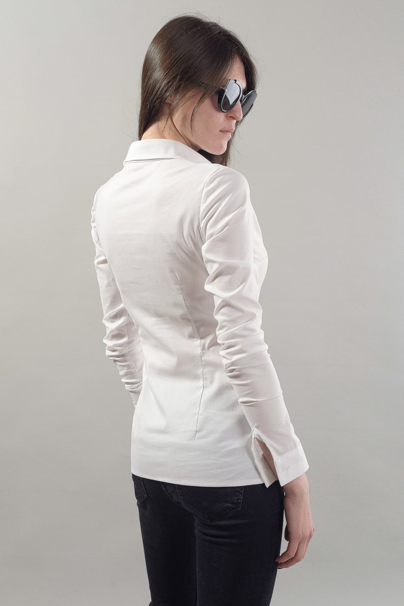 Puede incluir: Una camisa blanca de manga larga con botones y silueta ajustada. La camisa tiene cuello y mangas largas con pu&ntilde;os abotonados. La modelo lleva pantalones vaqueros negros.