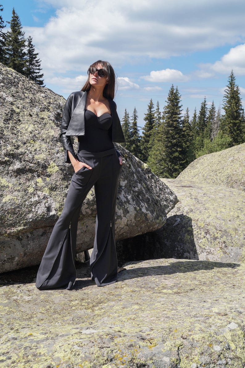 May include: A woman wearing a black leather jacket, a black bodysuit, and black flared pants with zippers on the sides. She is standing on a rocky surface with a forest in the background.