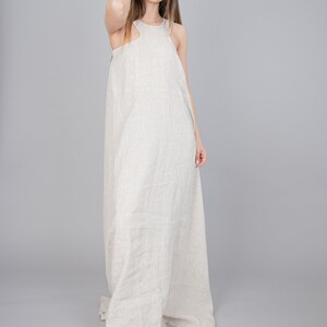 May include: A woman wearing a long, light beige linen dress with a sleeveless design. The dress has a relaxed fit and a flowing silhouette.