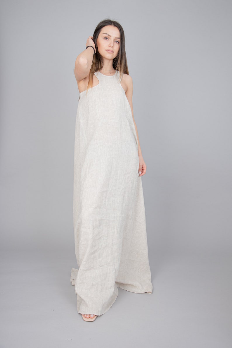 May include: A woman wearing a long, light beige linen dress with a sleeveless design. The dress has a relaxed fit and a flowing silhouette.