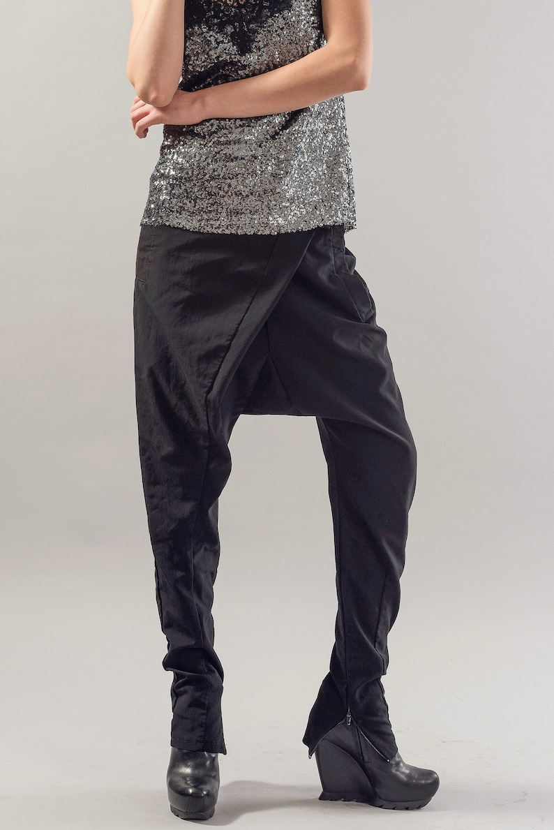 May include: Black trousers with a distinctive draped design and a wide leg silhouette. The trousers are made from a soft, flowing fabric and feature a zip detail at the ankle.