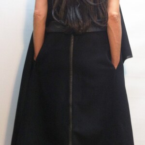 May include: A black sleeveless vest featuring a vertical leather-like trim down the centre back, leather-like hem detailing, and side pockets. The garment is labelled Flo Atelier and is worn over black trousers and black platform shoes.