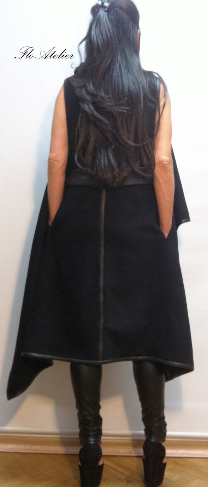 May include: A black sleeveless vest featuring a vertical leather-like trim down the centre back, leather-like hem detailing, and side pockets. The garment is labelled Flo Atelier and is worn over black trousers and black platform shoes.
