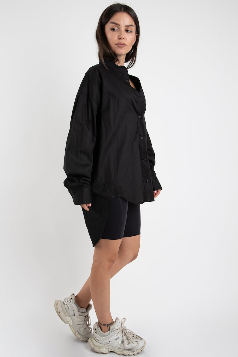 Puede incluir: Una camisa negra de botones de gran tama&ntilde;o con mangas largas y cuello alto. La camisa se lleva con pantalones cortos de ciclista negros y zapatillas blancas.