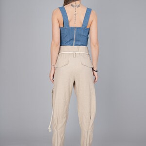 May include: A denim blue corset top with a silver zipper and beige linen cargo pants. The pants have a white belt and zippers at the ankles. The model is wearing light pink heels.