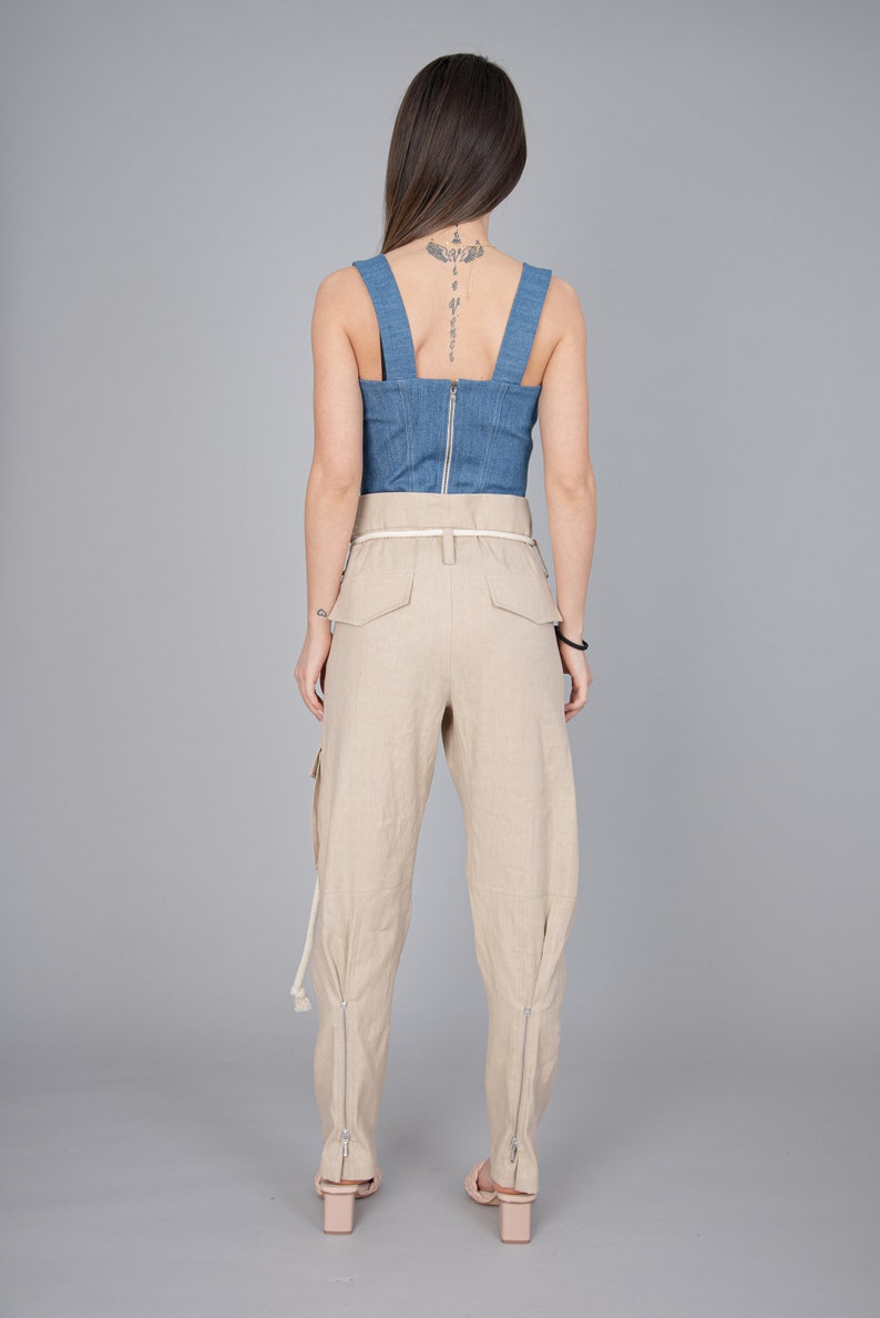 May include: A denim blue corset top with a silver zipper and beige linen cargo pants. The pants have a white belt and zippers at the ankles. The model is wearing light pink heels.