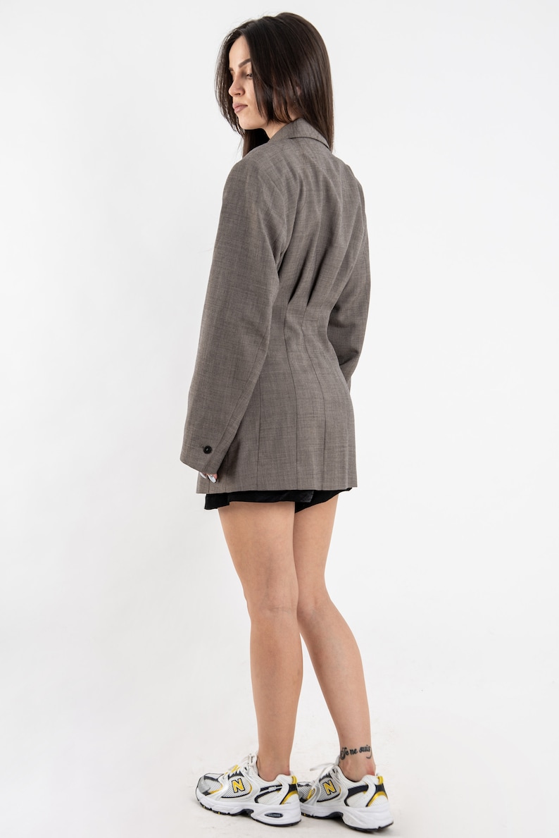 May include: A brown blazer with a fitted waist and long sleeves. The blazer has a single button closure and a notched collar. The model is wearing white sneakers with yellow accents.