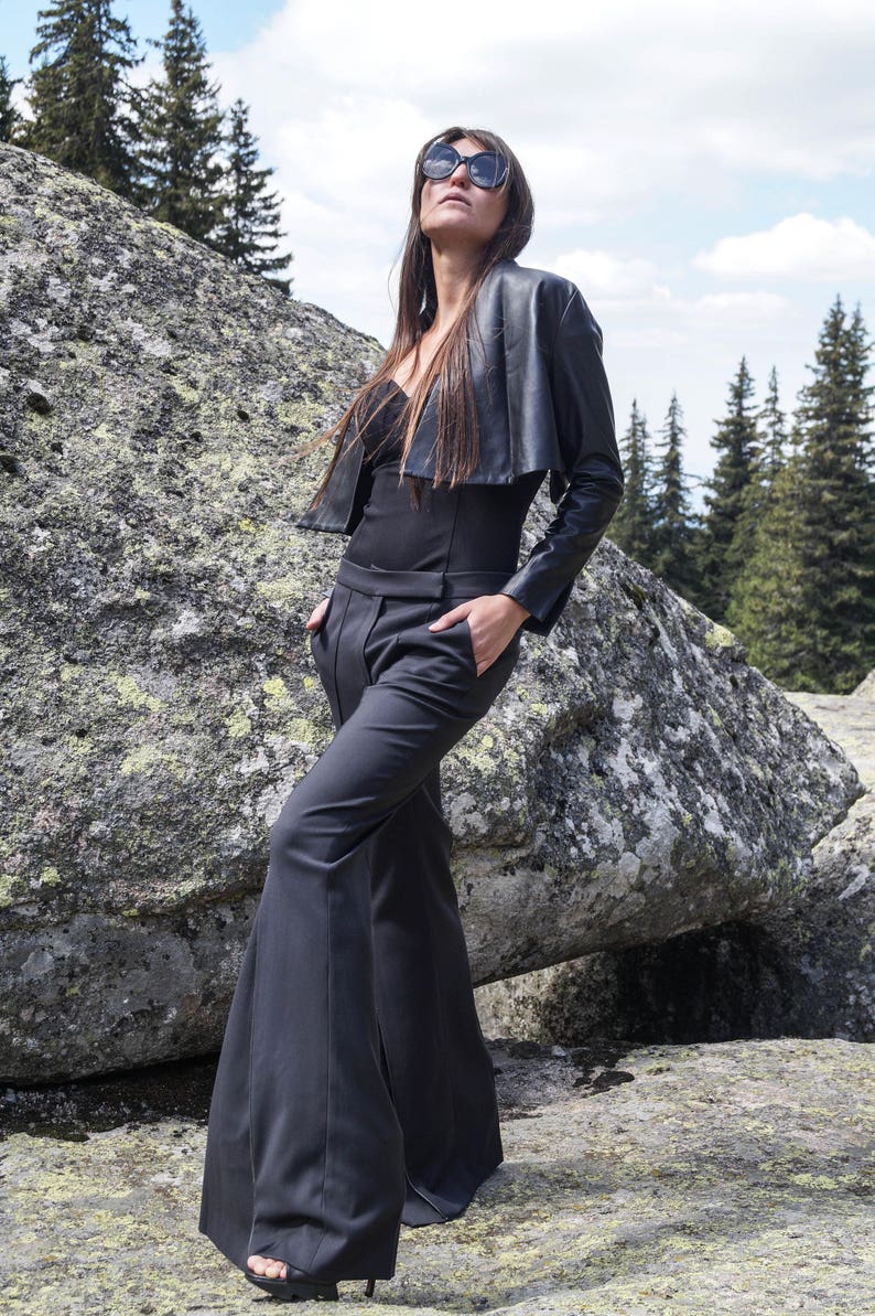May include: A woman wearing a black leather jacket, black pants, and sunglasses. She is standing in front of a large rock.