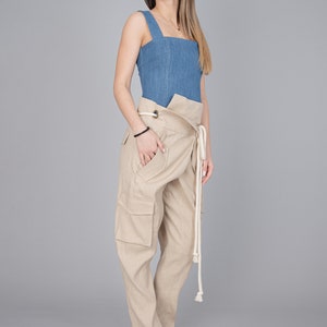 May include: A denim blue square-neck top paired with beige wrap-style cargo pants. The pants feature large pockets and a rope tie. The model is wearing beige woven heeled sandals. The outfit is set against a gray backdrop.