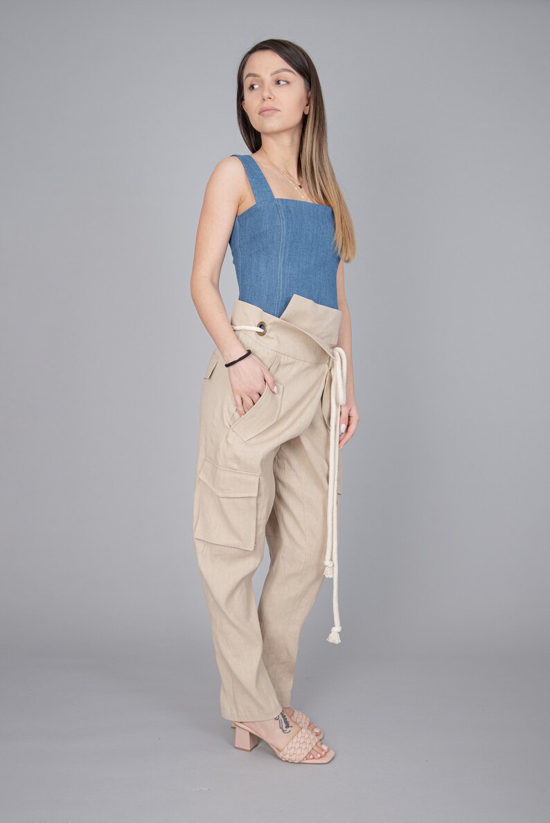 May include: A denim blue square-neck top paired with beige wrap-style cargo pants. The pants feature large pockets and a rope tie. The model is wearing beige woven heeled sandals. The outfit is set against a gray backdrop.