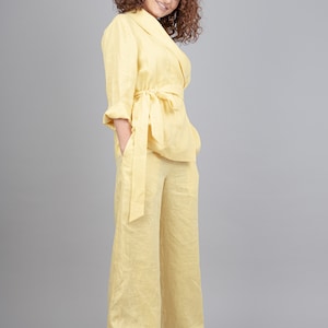 May include: A woman wearing a yellow linen pantsuit with a matching belt. She is standing with her hands in her pockets and smiling at the camera.