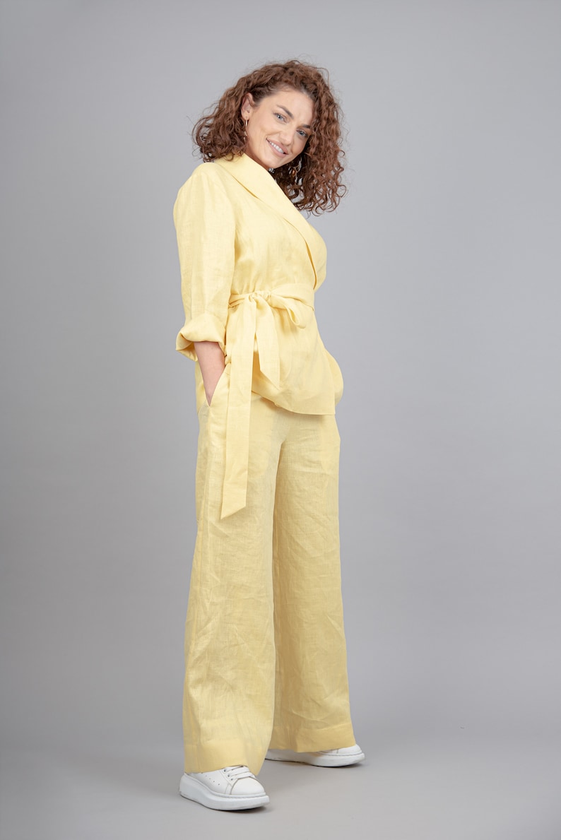 May include: A woman wearing a yellow linen pantsuit with a matching belt. She is standing with her hands in her pockets and smiling at the camera.