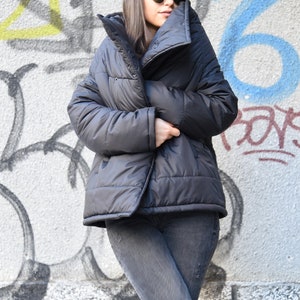 May include: A black puffer jacket with a high collar and a zipper closure. The jacket is worn by a person wearing a black cap and sunglasses.