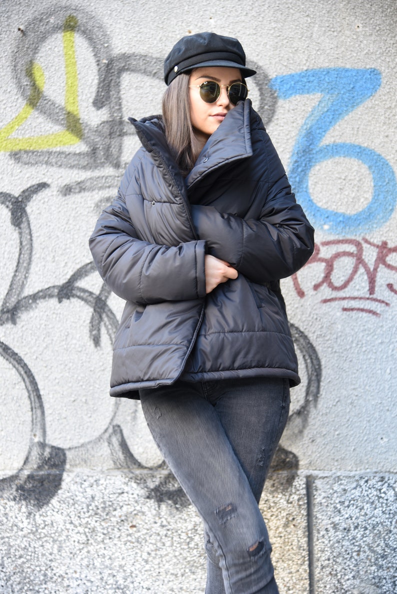 May include: A black puffer jacket with a high collar and a zipper closure. The jacket is worn by a person wearing a black cap and sunglasses.