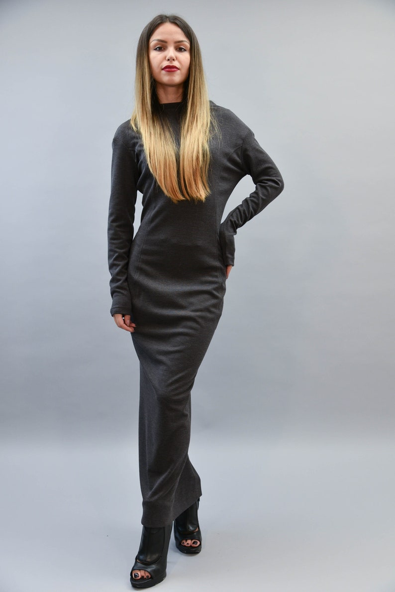 May include: A dark gray, long-sleeved maxi dress. The dress has a high neckline and a fitted silhouette. The model is wearing black open-toe boots. The background is a solid light gray.