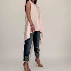 May include: A woman wearing a light pink sleeveless top with a high-low hemline and distressed blue jeans. She is wearing nude heels.