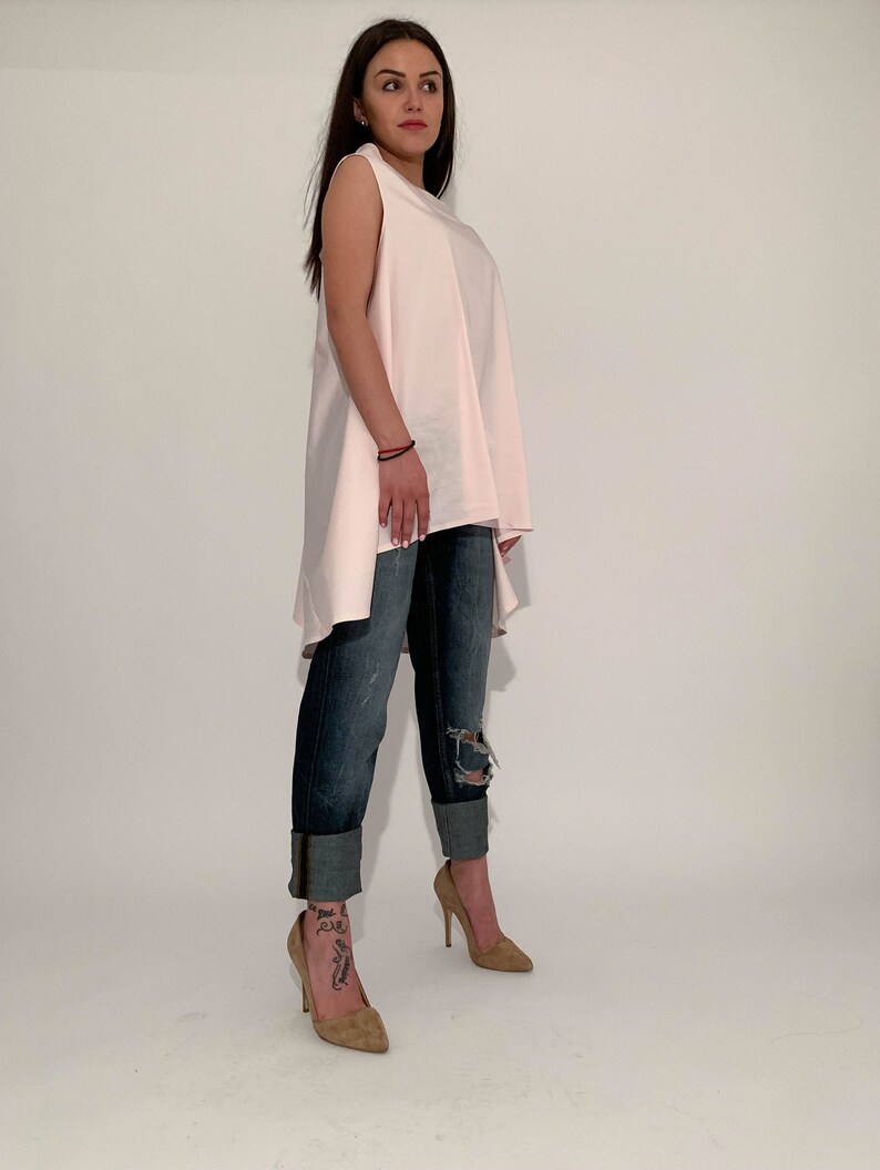 May include: A woman wearing a light pink sleeveless top with a high-low hemline and distressed blue jeans. She is wearing nude heels.