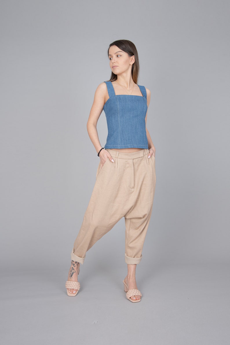 May include: A woman wearing a light blue denim tank top and beige, loose-fitting trousers. She is standing with her hands in her pockets and looking to the side.