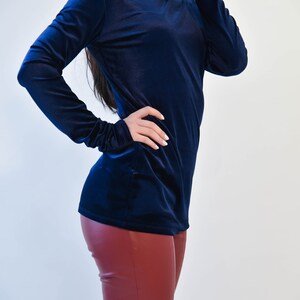 May include: A navy blue velvet turtleneck top with long sleeves. The top is fitted and has a smooth texture. The model is wearing the top with burgundy leather pants.