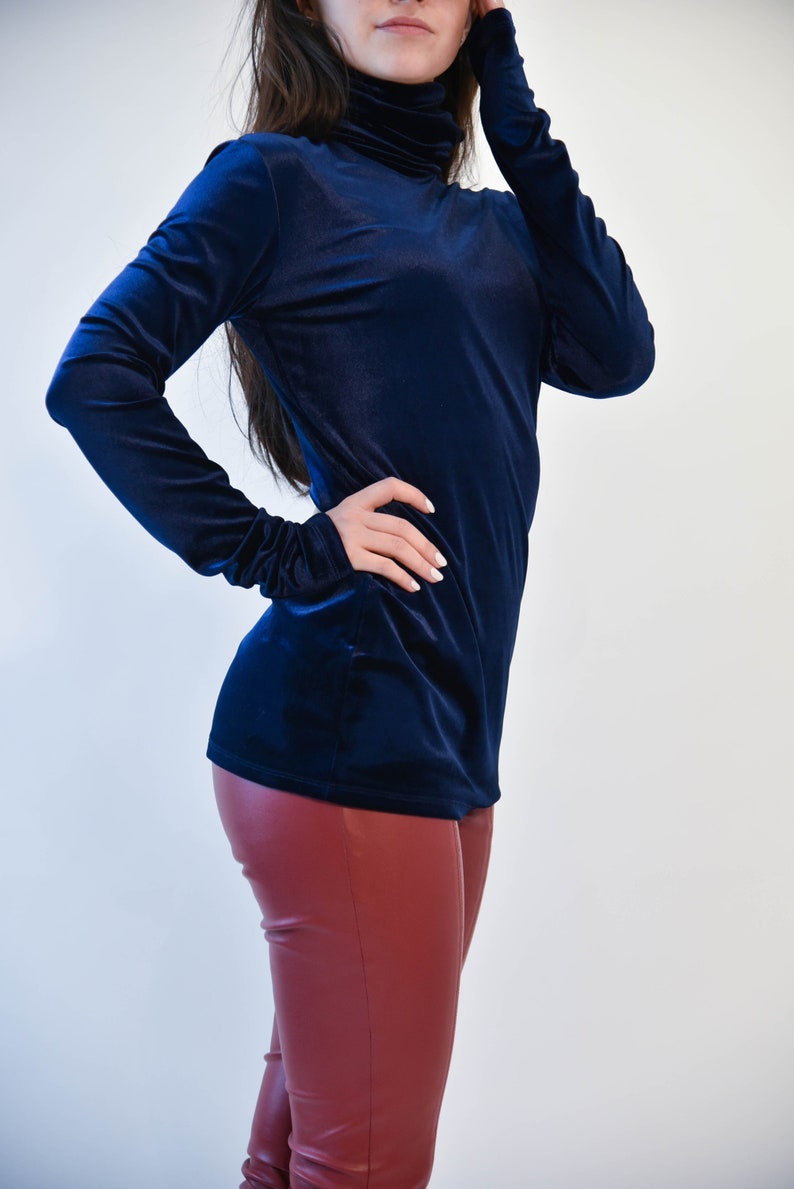 May include: A navy blue velvet turtleneck top with long sleeves. The top is fitted and has a smooth texture. The model is wearing the top with burgundy leather pants.