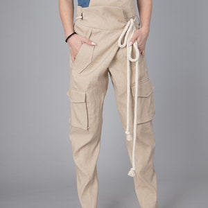 May include: Beige cargo pants with a wrap-around waist and a cream-colored rope belt. The pants have multiple pockets and are made of a light-colored fabric. The model is wearing a denim top and woven sandals.