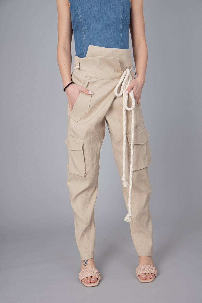 May include: Beige cargo pants with a wrap-around waist and a cream-colored rope belt. The pants have multiple pockets and are made of a light-colored fabric. The model is wearing a denim top and woven sandals.