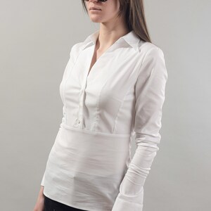 Puede incluir: Una camisa blanca de manga larga con botones y cuello en V. La camisa tiene una cintura ajustada y est&aacute; metida en un pantal&oacute;n negro.